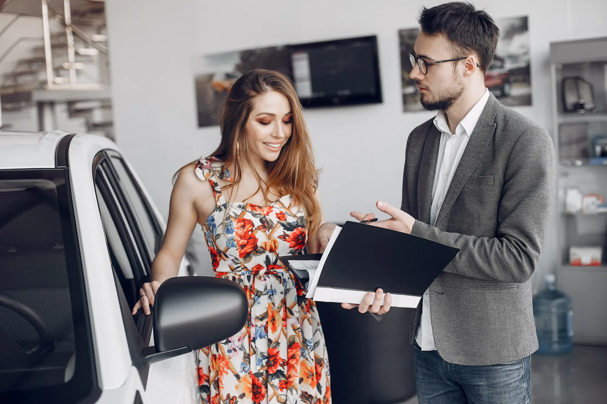 Car salesman in a suit showing documents to young woman in a automotive showroom with a white car in background